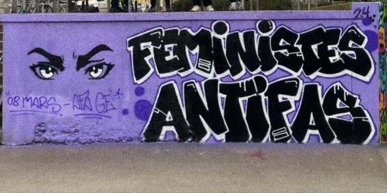 Bild einer Betonmauer in lila Flieder farben. Auf der rechten Seite zu sehen zwei Augen. Daneben steht als Graffiti in Schwarz. Feministes Antifas in schwarz.