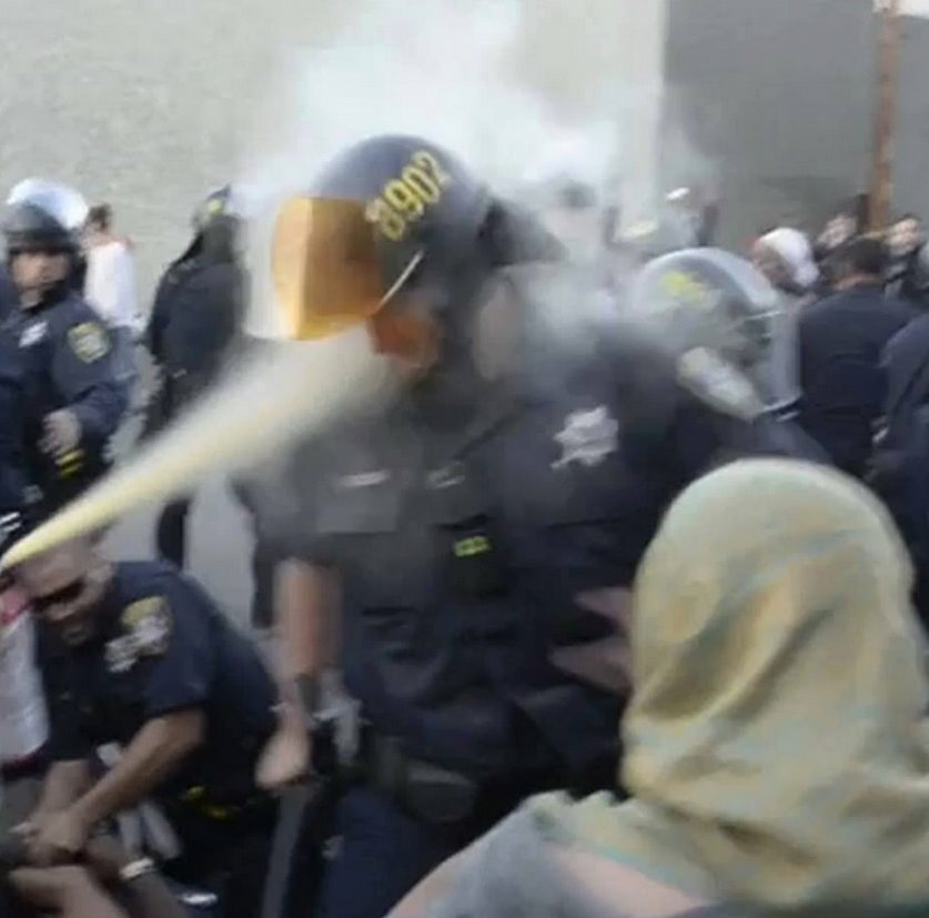 A blurred photo showing a riot cop being sprayed in the face with pepper spray by a demonstrator during a sit-in. There's a person with a headscarf in the foreground, several demonstrators and riot cops in the background.