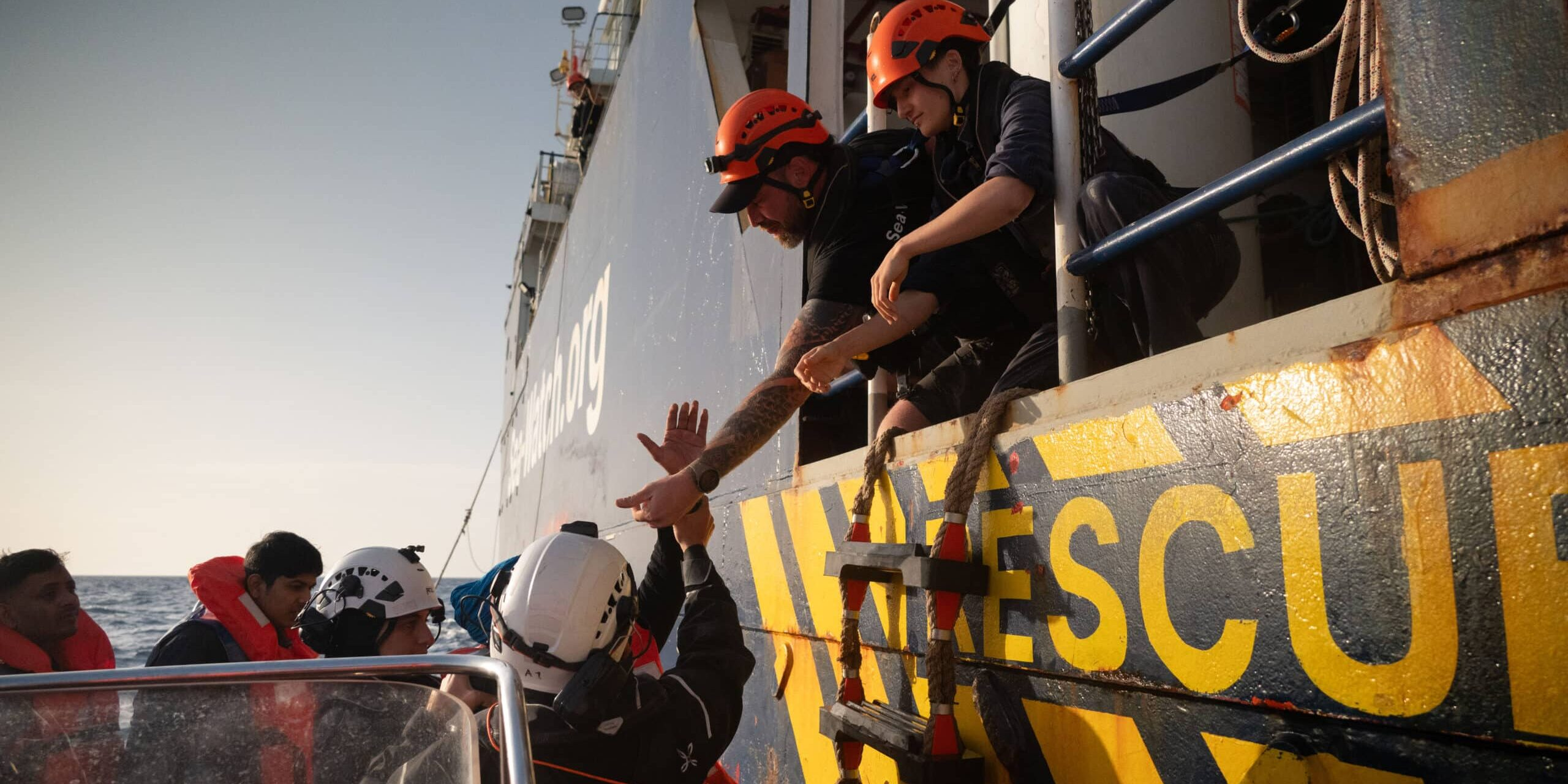 Zu sehen sind zwei Seenotretter*innen auf einem Schiff welche Menschen auf einem Boot die Hände reichen.
