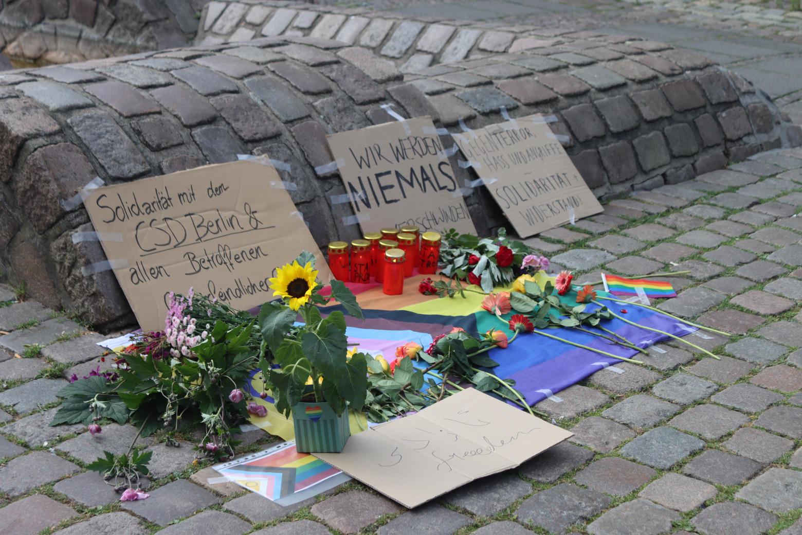 Auf einer progress pride flag liegen Blumen. Mehrere Schilder sind zu sehen. Auf einem Schild steht "Solidarität mit dem CSD Berlin und allen Betroffenen queerfeindlicher Gewalt". Auf einm anderen Schild steht "Wir werden niemals verschwinden."