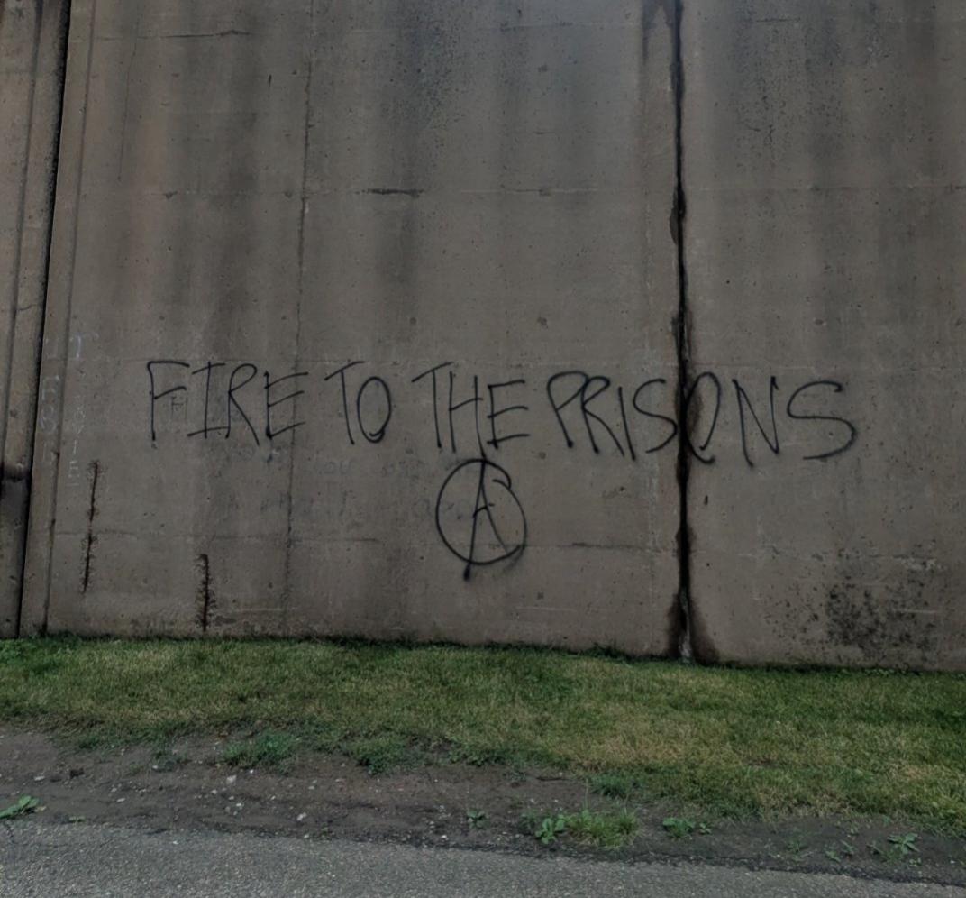 A gray wall by a roadside has "FIRE TO THE PRISONS Ⓐ" written on it in black ink