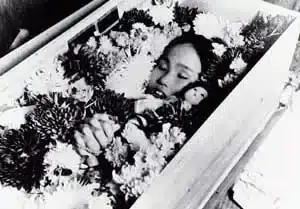 Ein schwarzweiß Foto: Die 12-jährige Sadako Sasaki liegt in ihrem Sarg . Zu sehen sind nur der Kopf des Mädchens und die Hände. Sie liegt in einem Meer aus Blumen. Sadako erkrankte nach dem Atombombenabwurf auf Hiroshima an Leukämie und starb 1955. Während ihres jahrelangen Krankenhausaufenthaltes faltete sie tausend Kraniche aus Origami-Papier.